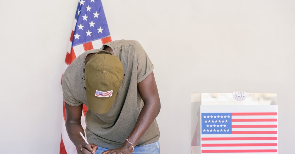 A voter in a cap fills out a ballot beside a ballot box and an American flag.